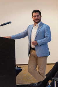 Un profesional de impecable presencia, ataviado con un blazer azul claro y pantalones caqui, se dirige a una audiencia desde el podio, exponiendo detalles clave sobre exclusivos proyectos residenciales. Su estilo sobrio y seguro -cabello oscuro cuidadosamente arreglado y barba bien cuidada- refleja el alto estándar propio del sector inmobiliario premium. La presentación, enfocada en propuestas habitacionales de alto valor, destaca por su claridad y orientación hacia inversionistas y compradores que buscan excelencia en ubicación, diseño arquitectónico e innovación. Al fondo, la ambientación neutra resalta la seriedad del evento e invita a concentrar la atención en los argumentos diferenciadores del portafolio presentado.