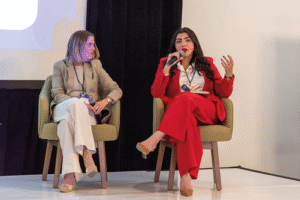 Dos mujeres se encuentran sentadas en el escenario, participando como ponentes destacadas. La oradora a la derecha, vestida con un elegante traje rojo, comparte su experiencia en sustentabilidad aplicada a proyectos de vivienda de alto estándar. Su intervención, respaldada por su actitud segura y su comunicación clara al micrófono, resalta las tendencias innovadoras que actualmente marcan pauta en el sector inmobiliario premium. Ambas portan credenciales de conferencistas, subrayando la relevancia y autoridad del evento para inversores y compradores exigentes interesados en propuestas residenciales exclusivas y sostenibles.