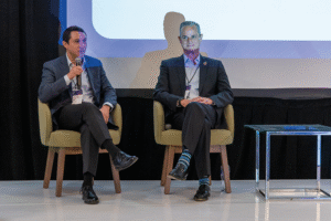 Dos ejecutivos de alto nivel, impecablemente vestidos, entablan un diálogo estratégico sobre comunidades planeadas desde el escenario principal. Sentados en elegantes butacas y con credenciales visibles que subrayan su relevancia en el sector, abordan temas clave para inversores y compradores sofisticados. Mientras uno expone con claridad los beneficios y ventajas competitivas de este tipo de desarrollos inmobiliarios premium a través de un micrófono, su interlocutor escucha atentamente, reflejando una visión analítica e informada. El entorno moderno incluye una mesa auxiliar de cristal y una pantalla como fondo, reforzando la atmósfera exclusiva y profesional del encuentro.