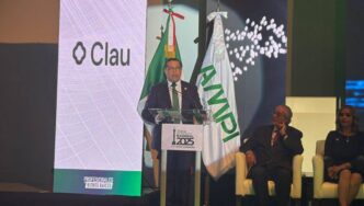 Un ejecutivo de alto nivel, vestido impecablemente, dirige su mensaje desde un podio institucional identificado con el Día Nacional 2025. El escenario está enmarcado por banderas oficiales de la AMPI y una imponente pantalla de gran formato que proyecta el logotipo de Clau. A su izquierda, dos distinguidos panelistas acompañan el evento, subrayando la relevancia y sofisticación del foro dirigido a líderes e inversionistas del sector inmobiliario premium.