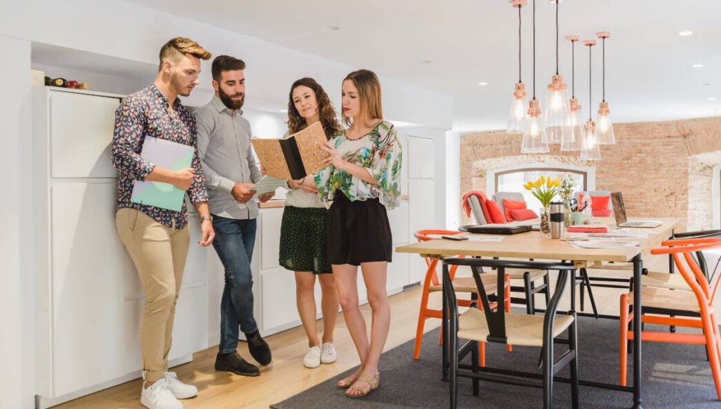 Cuatro jóvenes profesionales se reúnen en una cocina contemporánea y luminosa dentro de un exclusivo espacio de coliving en España. Una de ellas, con libreta en mano, guía la conversación mientras los demás participan activamente. En primer plano, una mesa de comedor elegante con sillas de diseño y arreglos florales resalta la atención al detalle y el ambiente cómodo que caracteriza a esta propiedad premium, pensada para quienes valoran comunidades vibrantes y espacios funcionales sin renunciar al lujo y la comodidad.