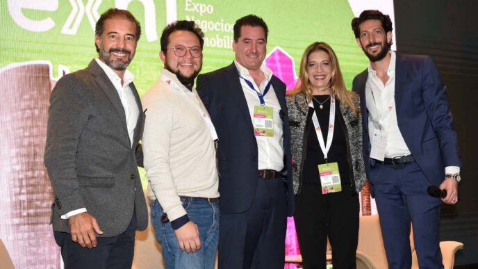 Cinco profesionales se posan juntos, mostrando una actitud cordial y segura durante un exclusivo evento de Renovación Propiedades. Todas lucen acreditaciones y presentan una imagen impecable con vestimenta smart casual, reflejando la elegancia propia del sector inmobiliario premium. Se ubican frente a un fondo cómodo en tonos verde y blanco con branding destacado de la firma. Uno de ellos sostiene un micrófono, listo para compartir su experiencia sobre oportunidades estratégicas y tendencias actuales del mercado inmobiliario de alta gama.