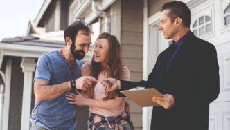 Un asesor inmobiliario entrega las llaves a una pareja frente a su nueva residencia, reflejando la satisfacción de una exitosa transacción en el sector residencial premium. Esta escena simboliza el cumplimiento de las altas expectativas de clientes exigentes, subrayando la excelencia en el servicio, la confianza y el acceso a propiedades exclusivas que distinguen al mercado inmobiliario de alto nivel.