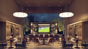 Exclusivo lounge en Origina Pedregal: un espacio contemporáneo con elegantes pisos de madera, mobiliario de diseño y mesas redondas que invitan a la convivencia. La iluminación suspendida crea una atmósfera sofisticada, complementada por una barra integrada de alta gama y una pantalla de gran formato para experiencias sociales premium. Ventanales panorámicas ofrecen vistas únicas a los jardines y al cielo nocturno, consolidando este ambiente como el punto de encuentro ideal para disfrutar y entretenerse en uno de los desarrollos residenciales más exclusivos del sur de la ciudad.