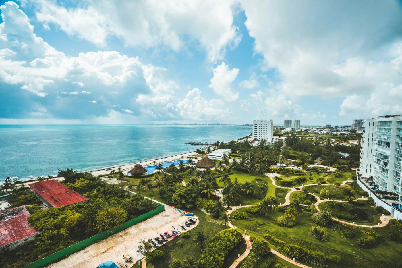 Vista aérea de una exclusiva ciudad costera en la Riviera Maya, donde imponentes edificios residenciales se integran armoniosamente con playas de arena blanca, albercas de diseño vanguardista y exuberantes áreas verdes. Destacan elegantes senderos entre palmeras y jardines meticulosamente cuidados, todo enmarcado por un cielo parcialmente nublado que resalta la belleza natural del entorno. Esta ubicación privilegiada representa una oportunidad única para inversionistas y compradores que buscan excelencia, privacidad y un estilo de vida cómodo frente al mar Caribe.