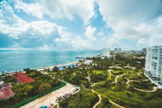 Vista aérea de una exclusiva ciudad costera en la Riviera Maya, donde imponentes edificios residenciales se integran armoniosamente con playas de arena blanca, albercas de diseño vanguardista y exuberantes áreas verdes. Destacan elegantes senderos entre palmeras y jardines meticulosamente cuidados, todo enmarcado por un cielo parcialmente nublado que resalta la belleza natural del entorno. Esta ubicación privilegiada representa una oportunidad única para inversionistas y compradores que buscan excelencia, privacidad y un estilo de vida cómodo frente al mar Caribe.