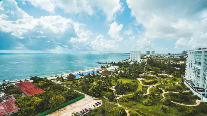 Vista aérea de una exclusiva ciudad costera en la Riviera Maya, donde imponentes edificios residenciales se integran armoniosamente con playas de arena blanca, albercas de diseño vanguardista y exuberantes áreas verdes. Destacan elegantes senderos entre palmeras y jardines meticulosamente cuidados, todo enmarcado por un cielo parcialmente nublado que resalta la belleza natural del entorno. Esta ubicación privilegiada representa una oportunidad única para inversionistas y compradores que buscan excelencia, privacidad y un estilo de vida cómodo frente al mar Caribe.