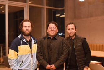 Tres ejecutivos se reúnen en un elegante espacio de WAS Company, caracterizado por amplios ventanales que inundan el ambiente de luz natural y un cómodo mobiliario en madera. La escena refleja un contexto de negocios premium: el primer socio viste chaqueta deportiva, el segundo opta por una presentación más formal con gafas y abrigo oscuro, mientras que el tercero apuesta por un chaleco negro moderno. Esta sala ejemplifica los altos estándares arquitectónicos y la atención al detalle que distingue a las propiedades exclusivas del mercado inmobiliario de lujo.