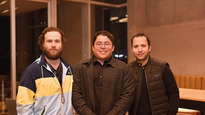 Tres ejecutivos se reúnen en un elegante espacio de WAS Company, caracterizado por amplios ventanales que inundan el ambiente de luz natural y un cómodo mobiliario en madera. La escena refleja un contexto de negocios premium: el primer socio viste chaqueta deportiva, el segundo opta por una presentación más formal con gafas y abrigo oscuro, mientras que el tercero apuesta por un chaleco negro moderno. Esta sala ejemplifica los altos estándares arquitectónicos y la atención al detalle que distingue a las propiedades exclusivas del mercado inmobiliario de lujo.