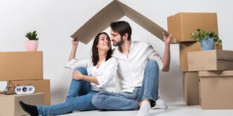 Una pareja celebra la adquisición de su nuevo hogar, rodeado de cajas de mudanza y detalles naturales que realzan el ambiente exclusivo de su residencia. Gracias al acceso a soluciones crediticias personalizadas, han hecho realidad su inversión en una propiedad premium. Juntos sostienen sobre sus cabezas una figura en forma de techo, símbolo del inicio de una nueva etapa marcada por seguridad, confianza y visión patrimonial en el sector inmobiliario de alta gama.