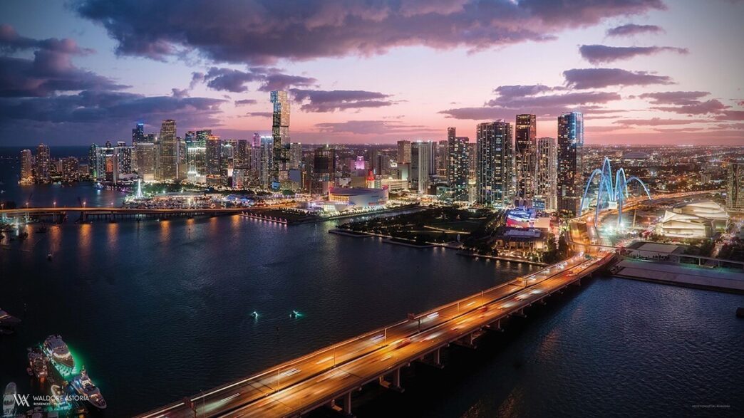Impresionante panorámica del centro de Miami al atardecer: rascacielos iluminados que definen el horizonte, elegantes puentes que cruzan las aguas y la vibrante luz urbana reflejándose en la Bahía de Biscayne. Un entorno cosmopolita y cómodo, ideal para quienes buscan invertir o residir en una de las zonas más exclusivas y dinámicas del sector inmobiliario premium.