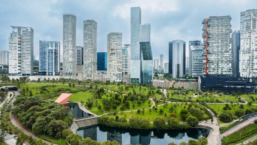 Panorama urbano de vanguardia con imponentes rascacielos modernos enmarcados por un parque arbolado, senderos peatonales, estanque y una exclusiva construcción de techo rojo. Este entorno, bajo un cielo matizado, refleja el desarrollo sostenible y la excelencia arquitectónica que caracterizan al sector inmobiliario premium. Inspirado por los más altos estándares de Holcim México, representa una oportunidad única para inversores y compradores exigentes que buscan integrar calidad de vida, innovación y plusvalía segura en espacios urbanos verdes y atractivos.