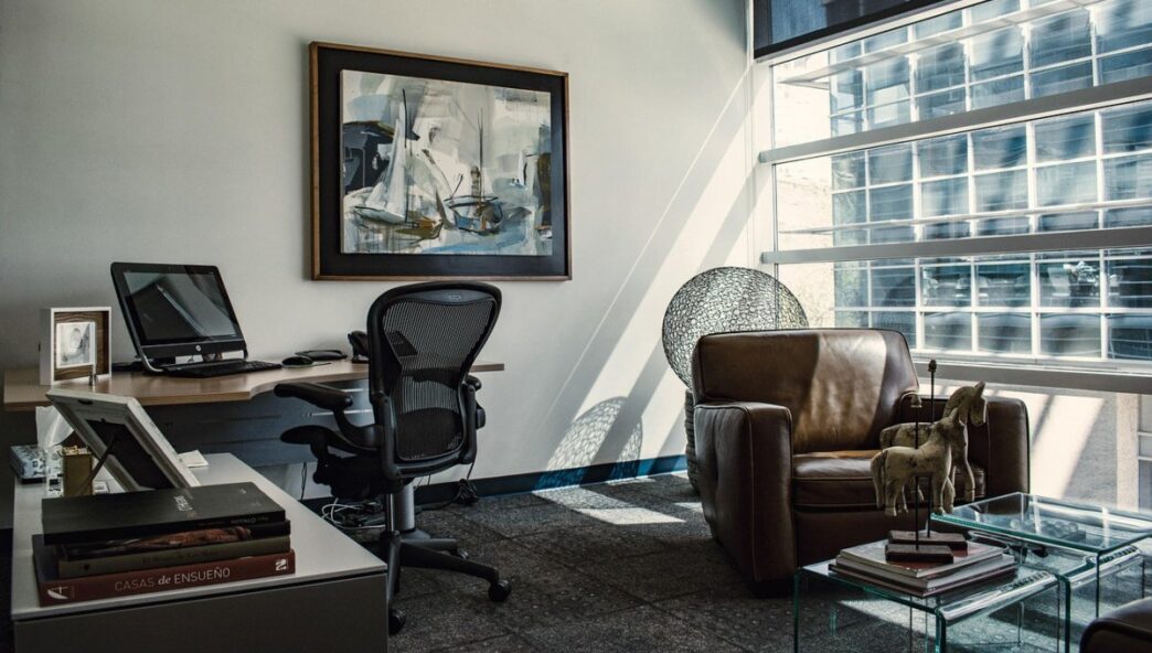 Oficina moderna equipada con mobiliario de alta gama: escritorio ejecutivo, ordenador de última generación y silla ergonómica, complementados por un elegante sillón de cuero junto a amplios ventanales que inundan el espacio de luz natural. Una obra abstracta marcada aporta sofisticación al ambiente, reflejando el diseño contemporáneo característico de las oficinas premium gestionadas por CBRE. Un entorno pensado para satisfacer las expectativas de inversores y compradores exigentes que valoran calidad, funcionalidad y prestigio en cada detalle.