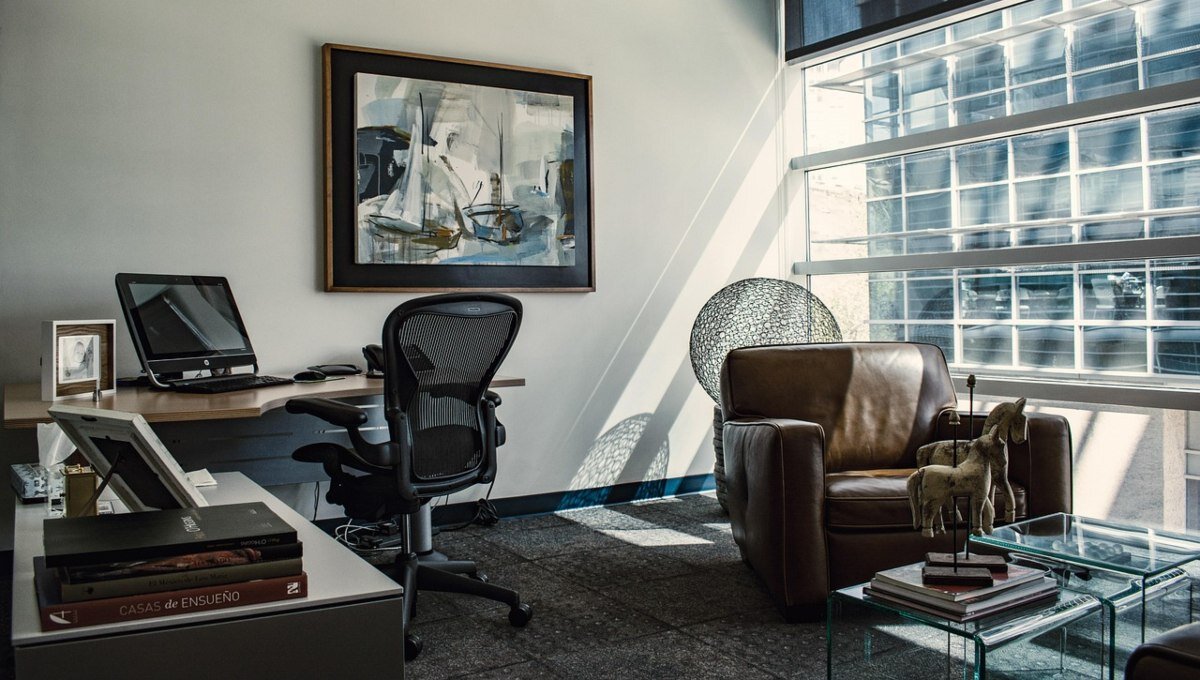 Oficina moderna equipada con mobiliario de alta gama: escritorio ejecutivo, ordenador de última generación y silla ergonómica, complementados por un elegante sillón de cuero junto a amplios ventanales que inundan el espacio de luz natural. Una obra abstracta marcada aporta sofisticación al ambiente, reflejando el diseño contemporáneo característico de las oficinas premium gestionadas por CBRE. Un entorno pensado para satisfacer las expectativas de inversores y compradores exigentes que valoran calidad, funcionalidad y prestigio en cada detalle.