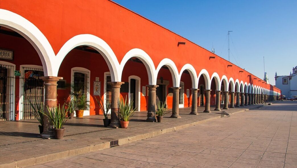 Un majestuoso edificio rojo de líneas alargadas destaca por sus elegantes arcos blancos y columnas de piedra, evocando la arquitectura tradicional poblana con un toque contemporáneo. Esta propiedad exclusiva bordea una plaza pavimentada, creando un entorno cómodo e inspirador. La presencia de plantas ornamentales cuidadosamente dispuestas entre las columnas y la vista despejada hacia un cielo azul completan una atmósfera inigualable, ideal para inversionistas o compradores que buscan autenticidad arquitectónica, distinción y alto potencial de valorización en el mercado inmobiliario premium.