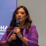 Una conferencista de alto perfil, reconocida por su experiencia en el sector inmobiliario de lujo, exponente sobre innovación tecnológica en bienes raíces (proptech). Vestida elegantemente con un blazer púrpura, comparte sus conocimientos y perspectivas ante una audiencia selecta e inversores exigentes. El evento se desarrolla en un entorno corporativo exclusivo, acompañado del emblema de Wyndham Hotels & Resorts como referente global de excelencia y prestigio en hospitalidad.