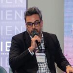 Un ejecutivo de amplia trayectoria en el sector inmobiliario premium, distinguido por su cabello canoso y gafas, viste un blazer negro y una camisa a cuadros mientras interviene como ponente ante un público selecto. Porta una credencial oficial, reflejo de su participación en un evento exclusivo organizado por CIEN Grupo Inmobiliario. Al fondo, se exhibe un banner corporativo que resalta los avances más innovadores en proptech e impulsa la visión de vanguardia del grupo; junto a este, una pantalla proyecta información relevante para inversores y compradores exigentes interesados en oportunidades diferenciadas dentro del mercado inmobiliario de alto valor.