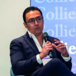 Un distinguido ponente, ataviado con un elegante traje azul marino y gafas, exponen sobre temas de geopolítica durante un evento exclusivo del sector inmobiliario. Su credencial de identificación resalta su participación como orador principal. El escenario cuenta con una sofisticada señalización corporativa en azul, destacando la marca Colliers, símbolo de excelencia y liderazgo en el mercado inmobiliario premium.