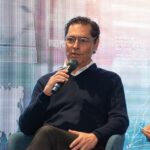 Un distinguido ponente, reconocido por su experiencia en geopolítica, participa como conferenciante principal en un evento exclusivo. Ataviado con indumentaria elegante y moderna, sostiene el micrófono mientras comparte perspectivas estratégicas ante una audiencia selecta. El fondo, compuesto por un mapa mundial artístico y elementos abstractos de diseño contemporáneo, refuerza la atmósfera global e innovadora del encuentro. Esta escena ejemplifica el nivel de sofisticación y visión internacional que caracteriza a las experiencias asociadas con los activos inmobiliarios premium: espacios donde convergen líderes influyentes para debatir tendencias que impactan el mercado mundial y generan oportunidades de inversión excepcionales.