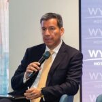 Un ejecutivo de alto nivel, vestido con un elegante traje oscuro y corbata amarilla, se dirige a una audiencia especializada mientras analiza temas de geopolítica con visión estratégica. Sentado con porte profesional, sostiene un micrófono en la mano y toma notas cuidadosamente, demostrando su enfoque analítico. De fondo sobresale el emblemático logo de Wyndham Hotels & Resorts, sinónimo de excelencia y liderazgo global en hospitalidad premium, un contexto ideal para inversores y compradores que buscan oportunidades exclusivas en el mercado inmobiliario internacional.