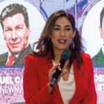 Una mujer con blazer rojo toma la palabra frente al auditorio, transmitiendo profesionalismo y liderazgo. Al fondo, dos destacadas imágenes circulares exhiben a Samuel y José Juven en pantalla, anticipando un análisis de alto nivel sobre geopolítica. Este contexto dota al evento de un carácter exclusivo e intelectual, alineado con los estándares de quienes buscan invertir o adquirir propiedades en el sector inmobiliario premium: líderes que valoran visión global, información privilegiada y entornos favorables para tomar decisiones estratégicas.