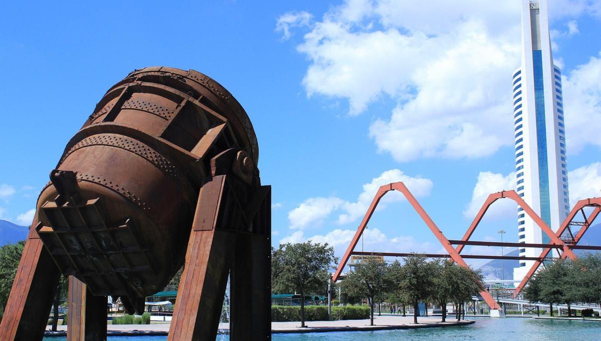 Una imponente escultura industrial de tono óxido se erige sobre una plataforma junto al canal, en una de las ubicaciones más privilegiadas de Nuevo León. Al fondo, la silueta vanguardista de un rascacielos blanco y la arquitectura singular de un puente naranja en arco subrayan el dinamismo urbano y el desarrollo sofisticado que caracterizan a este enclave. Todo ello, enmarcado por un cielo azul con nubes dispersas, ofrece la perspectiva ideal para inversionistas y compradores que buscan integración única entre arte contemporáneo, infraestructura modernista y exclusividad inmobiliaria.