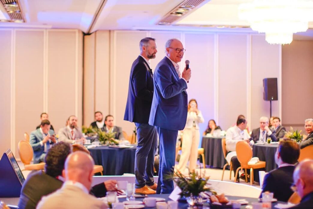 En el marco de un exclusivo evento organizado por Cushman & Wakefield, dos destacados profesionales del sector se posicionan en un elegante escenario. Uno de ellos, micrófono en mano, se dirige a una audiencia selecta compuesta por inversionistas y líderes del mercado inmobiliario premium, reunidos en un cómodo salón iluminado con calidez y dispuestos en mesas redondas. Este ambiente refleja la excelencia y distinción que caracteriza a los encuentros de alto nivel dedicados a las oportunidades más atractivas e innovadoras dentro del real estate de lujo.