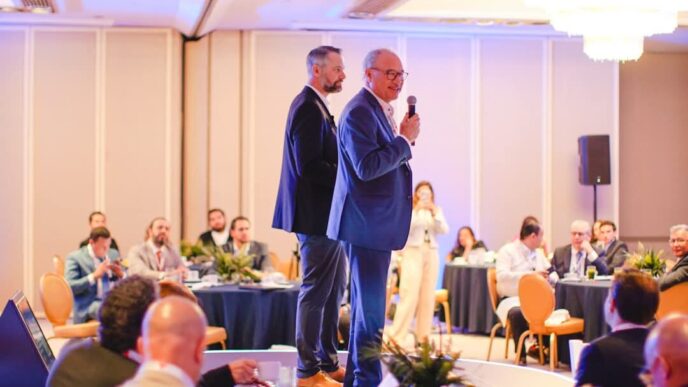 En el marco de un exclusivo evento organizado por Cushman & Wakefield, dos destacados profesionales del sector se posicionan en un elegante escenario. Uno de ellos, micrófono en mano, se dirige a una audiencia selecta compuesta por inversionistas y líderes del mercado inmobiliario premium, reunidos en un cómodo salón iluminado con calidez y dispuestos en mesas redondas. Este ambiente refleja la excelencia y distinción que caracteriza a los encuentros de alto nivel dedicados a las oportunidades más atractivas e innovadoras dentro del real estate de lujo.