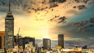Panorámica urbana al atardecer que revela un horizonte cómodo, dominado por imponentes rascacielos y una torre emblemática al lado izquierdo. El horizonte, enmarcado por nubes dispersas y la luz dorada del sol filtrándose entre dos edificios principales, resalta el dinamismo y la exclusividad de una ciudad cosmopolita ideal para inversiones inmobiliarias premium.