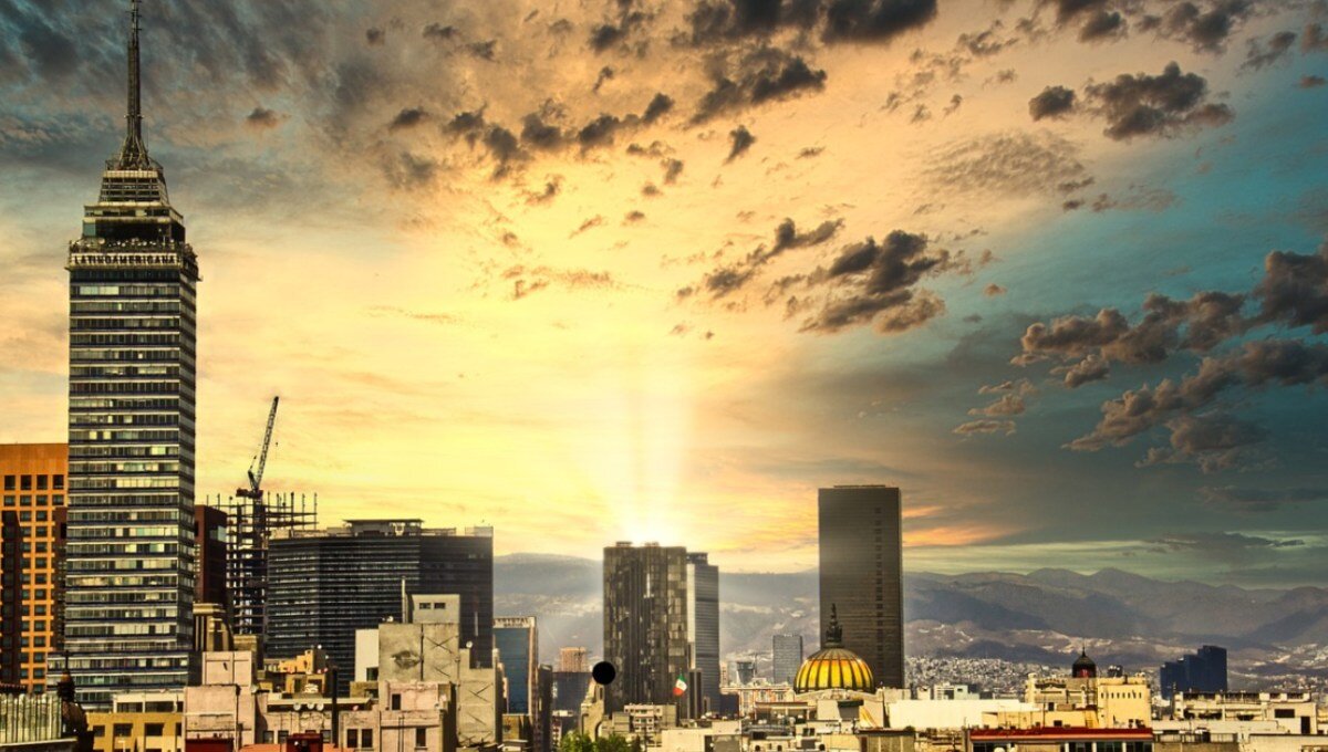 Panorámica urbana al atardecer que revela un horizonte cómodo, dominado por imponentes rascacielos y una torre emblemática al lado izquierdo. El horizonte, enmarcado por nubes dispersas y la luz dorada del sol filtrándose entre dos edificios principales, resalta el dinamismo y la exclusividad de una ciudad cosmopolita ideal para inversiones inmobiliarias premium.