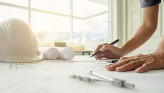 Un profesional revisa detalladamente planos arquitectónicos sobre su escritorio, utilizando instrumentos de precisión como un compás y una pluma. En la superficie destacan un casco de seguridad blanco y planos enrollados, reflejando el compromiso con los más altos estándares en diseño y construcción premium. La abundante luz natural que ingresa por los amplios ventanales acentúa la modernidad y el enfoque en espacios luminosos propios del sector inmobiliario de alto nivel. Escenarios como este son características en proyectos distinguidos, posibles candidatos a reconocimientos internacionales como los premios IWBI 2024, donde la excelencia en innovación y sostenibilidad es prioridad para inversionistas y compradores exigentes.