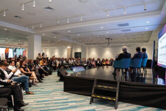 Un panel de expertos inmobiliarios se encuentra en el escenario, dirigiéndose a una audiencia selecta en un cómodo salón de conferencias. Los asistentes, atentos y comprometidos—muchos tomando notas y capturando imágenes clave—demuestran un interés genuino por las tendencias más exclusivas del sector. Una pantalla de gran formato acompaña la presentación, ofreciendo información relevante y actualizada sobre oportunidades de inversión premium, dinámicas del mercado y desarrollos emblemáticos para compradores e inversores que buscan excelencia y valor agregado en bienes raíces de alto nivel.