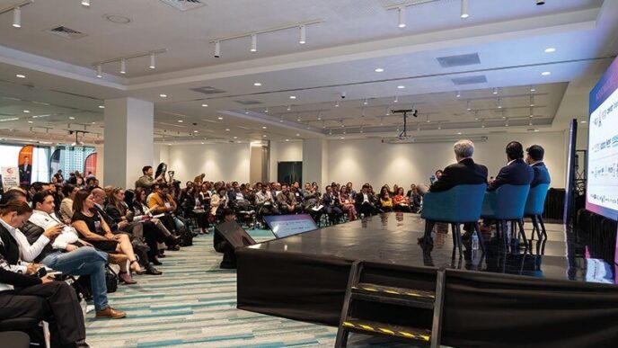 Un panel de expertos inmobiliarios se encuentra en el escenario, dirigiéndose a una audiencia selecta en un cómodo salón de conferencias. Los asistentes, atentos y comprometidos—muchos tomando notas y capturando imágenes clave—demuestran un interés genuino por las tendencias más exclusivas del sector. Una pantalla de gran formato acompaña la presentación, ofreciendo información relevante y actualizada sobre oportunidades de inversión premium, dinámicas del mercado y desarrollos emblemáticos para compradores e inversores que buscan excelencia y valor agregado en bienes raíces de alto nivel.