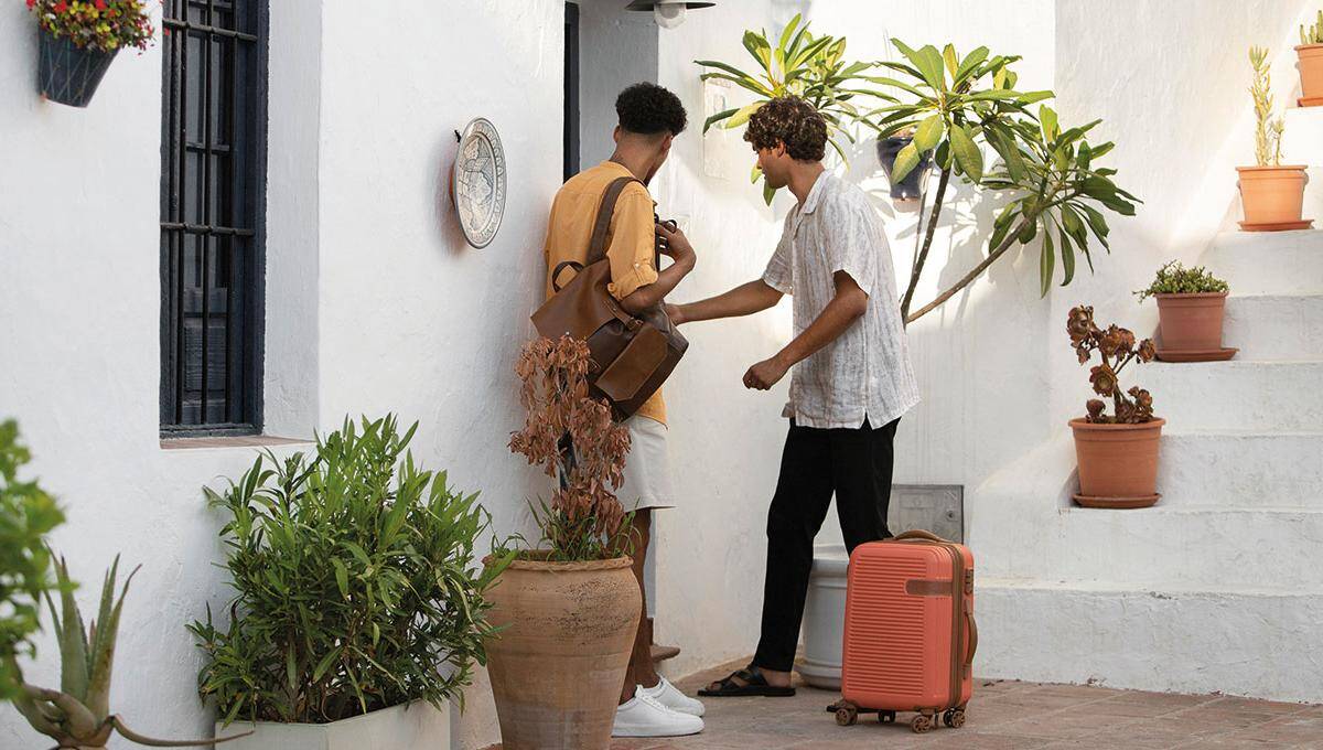 Dos personas se encuentran frente a una exclusiva fachada blanca, embellecida con elegantes plantas en maceta. Una de ellas accede al interior utilizando una llave y transportando una maleta de viaje, evocando la sofisticación y hospitalidad propia de los desarrollos inmobiliarios premium. La otra, con bolso al hombro, complementa este entorno distinguido que responde a las expectativas de compradores e inversores que valoran el diseño superior y la experiencia residencial de alto nivel.