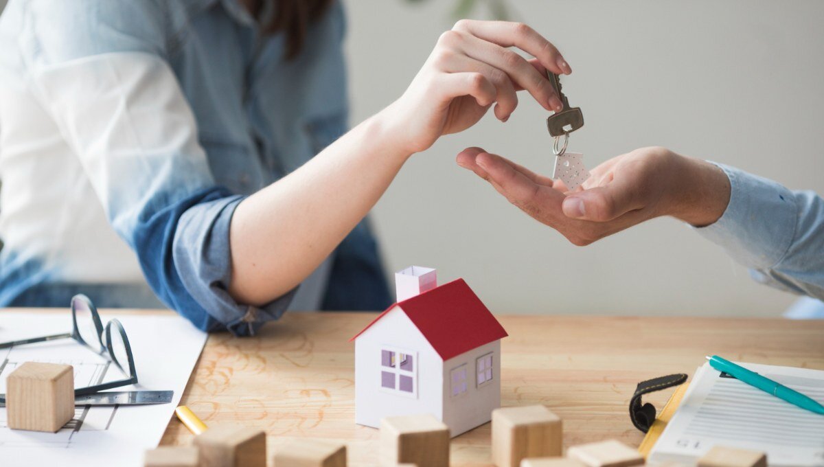 Una persona entrega la llave de una propiedad a otra, sobre una mesa con documentación, una maqueta residencial y bloques de madera. Esta escena representa el dinámico mercado inmobiliario premium en México, donde la activa demanda por inmuebles de alta gama evidencia el atractivo y solidez del sector para inversores y compradores beneficiosos que buscan activos exclusivos con alto potencial de valorización.