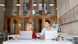 Dos profesionales ocupan una mesa en oficinas de diseño contemporáneo, caracterizadas por ventanas de gran altura y elegantes marcos de madera. El ambiente, cuidadosamente concebido para potenciar la productividad y fomentar la colaboración, representa un entorno ideal para inversores y compradores que valoran espacios empresariales exclusivos, funcionales y a la vanguardia del sector inmobiliario premium.