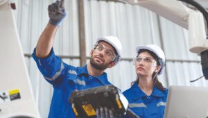 Dos profesionales especializados, equipados con uniformes corporativos y elementos de seguridad como cascos y gafas protectoras, inspeccionan avanzados sistemas tecnológicos en interiores. Uno de ellos indica un punto estratégico mientras opera un dispositivo de control, demostrando la integración de tecnología innovadora que optimiza los estándares de seguridad y eficiencia en entornos inmobiliarios premium. Este enfoque garantiza a inversores y compradores espacios exigentes con las mejores prácticas en protección, operatividad e infraestructura de alto nivel.