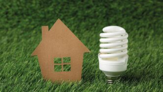 Un recorte representativo de una vivienda y una lámpara de bajo consumo se sitúan sobre césped natural, evocando el concepto de vivienda sustentable. Esta imagen transmite un firme compromiso con la eficiencia energética y la responsabilidad ambiental, elementos esenciales en el segmento inmobiliario premium para inversores y compradores que priorizan la innovación, la sostenibilidad y el valor a largo plazo.