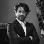 Un hombre de cabello oscuro a la altura media y barba cuidadosamente recortada, vestido con un elegante traje oscuro y camisa blanca impecable, posa con los brazos cruzados y una expresión segura dirigida a la cámara. Al fondo, el sutil diseño revela detalles del exclusivo entorno de residencias de marca en un enclave turístico premium en Baja California, reflejando el nivel superior de sofisticación y distinción que buscan inversionistas y compradores exigentes en propiedades de lujo.