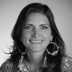 Imagen en blanco y negro de una mujer con larga cabellera oscura, que transmite confianza y sofisticación a través de su sonrisa. Luce pendientes circulares de gran tamaño y una blusa de diseño exclusivo inspirado en el estilo distintivo de Baja. El fondo, neutro y desenfocado, dirige toda la atención hacia la protagonista, resaltando su presencia magnética—un reflejo del carácter único que distingue a quienes buscan propiedades premium en destinos selectos.