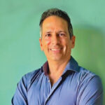 Un hombre de tez bronceada y cabello oscuro posa frente a un fondo teal, proyectando una actitud relajada y sofisticada acorde al exclusivo estilo de vida turística de Baja. Vestido con una camisa azul abotonada, su porte y confianza reflejan los valores fundamentales del sector inmobiliario premium: distinción, calidez y una proximidad auténtica al lujo. Una invitación visual ideal para inversionistas y compradores exigentes que buscan propiedades excepcionales en destinos de alta demanda.