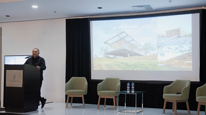 Un conferenciante especializado se presenta ante un selecto auditorio en un moderno salón de conferencias. En el escenario, el mobiliario minimalista -tres sillas exclusivas y una mesa con agua premium- subraya la atención al detalle y la calidad del entorno. La gran pantalla central proyecta información e imágenes sobre "Arquitectura del Nuevo Siglo", destacando innovaciones en diseño, sostenibilidad y lujo. Esta presentación está dirigida a inversores y compradores exigentes que buscan oportunidades únicas en desarrollos inmobiliarios de alto nivel, donde la excelencia arquitectónica se fusiona con el estilo de vida sofisticado.