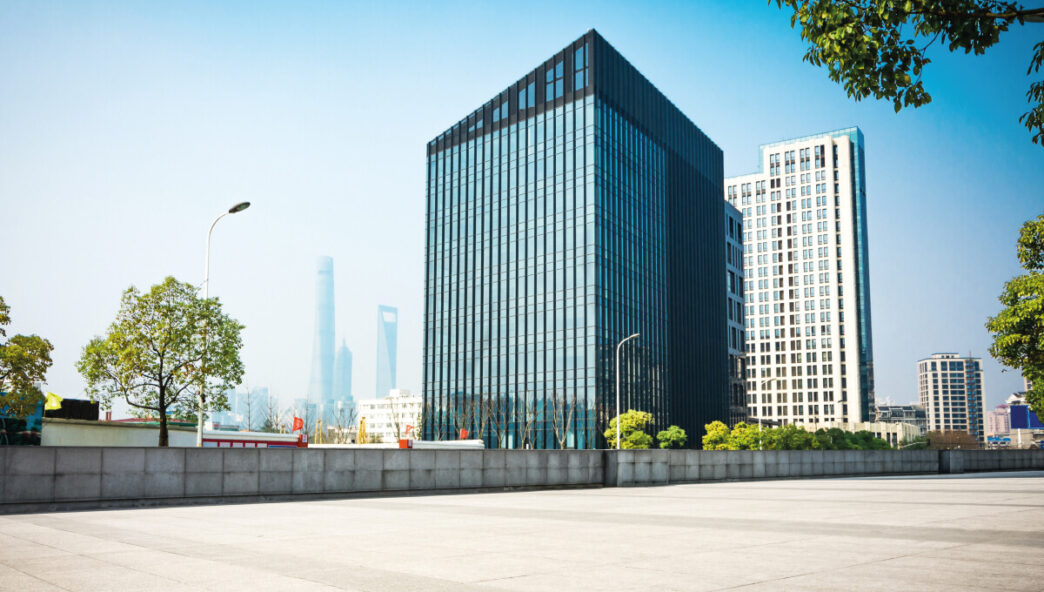 Un imponente edificio corporativo de diseño contemporáneo, con fachada de vidrio, se erige junto a una elegante torre blanca que evoca el estilo exclusivo de las residencias de marca en Baja. La plaza, perfectamente urbanizada y bordeada por árboles maduros, ofrece un ambiente sereno y cómodo. Al fondo, se perfilan rascacielos bajo un cielo despejado, completando un entorno urbano ideal para inversores o compradores que buscan propiedades premium en ubicaciones emblemáticas con proyección de alta plusvalía.