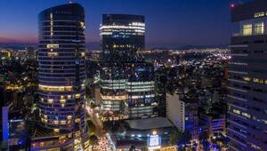 Panorama urbano al anochecer en 2030, donde modernos rascacielos de líneas elegantes se destacan por sus fachadas iluminadas y ventanales acristalados con vistas espectaculares. La ciudad cobra vida bajo un cielo azul profundo, mientras las calles brillan con la luz sofisticada de los vehículos y el alumbrado público. Este entorno vanguardista refleja una apuesta decidida por la sostenibilidad y la integración de tecnologías inteligentes, creando un destino ideal para inversores y compradores que buscan exclusividad, innovación y valor patrimonial a largo plazo en el sector inmobiliario premium.