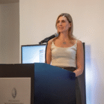 Una mujer, elegantemente vestida con un top blanco sin mangas, toma la palabra en el podio equipado con micrófono, presentando exclusivas oportunidades de inversión inmobiliaria premium ante una audiencia selecta. El entorno, sobrio y sofisticado, se realza con la presencia del logotipo de REBS Puebla y el emblema del Presidente InterContinental en un monitor cercano, reflejando el respaldo de marcas de prestigio internacional. Esta presentación subraya el acceso a activos inmobiliarios únicos y de alto valor estratégico para inversores visionarios y compradores exigentes que buscan lo mejor del mercado mexicano.