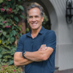 Un hombre de cabello castaño corto, vestido con un polo azul marino y un smartwatch, posa al aire libre con los brazos cruzados y una expresión confiada. Al fondo, la vegetación exuberante enmarca una exclusiva residencia de marca de tonos claros, reflejando el estándar más alto de lujo turístico en Baja. Esta imagen representa la sofisticación y el estilo de vida premium que buscan los inversores y compradores exigentes: privacidad, diseño contemporáneo y ubicación privilegiada en uno de los destinos más codiciados del mercado inmobiliario.