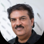 Un hombre de mediana edad, cabello oscuro y bigote bien cuidado, luce una camisa negra de corte impecable mientras dirige una ligera sonrisa a la cámara. El fondo luminoso y desenfocado, con sutiles patrones geométricos, refleja la sofisticación contemporánea característica de las residencias de marca en Baja. Esta imagen transmite el estilo refinado y exclusivo que buscan los inversores y compradores exigentes en propiedades inmobiliarias premium del destino.