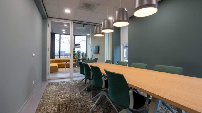 La sala de juntas, de diseño contemporáneo, destaca por su amplia mesa de madera, elegantes sillas verdes y luminarias colgantes que aportan sofisticación al espacio. Este ambiente ejemplifica la recuperación del sector inmobiliario corporativo premium en México rumbo a 2025. Las puertas de cristal dan paso a una exclusiva zona lounge con sofás amarillos y ventanas de gran formato, ofreciendo un entorno luminoso e inspirador que responde a las más altas expectativas de inversionistas y compradores exigentes.