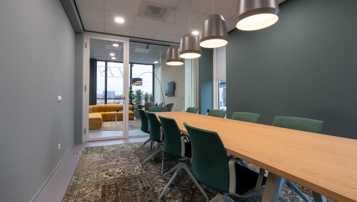 La sala de juntas, de diseño contemporáneo, destaca por su amplia mesa de madera, elegantes sillas verdes y luminarias colgantes que aportan sofisticación al espacio. Este ambiente ejemplifica la recuperación del sector inmobiliario corporativo premium en México rumbo a 2025. Las puertas de cristal dan paso a una exclusiva zona lounge con sofás amarillos y ventanas de gran formato, ofreciendo un entorno luminoso e inspirador que responde a las más altas expectativas de inversionistas y compradores exigentes.