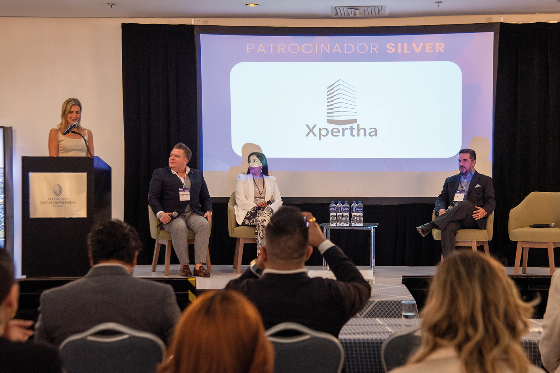 En el marco del exclusivo congreso REBS Puebla, cuatro destacados profesionales del sector inmobiliario se presentan en el escenario: uno de ellos lidera la conversación desde el podio, mientras los otros tres expertos acompañan la sesión como panelistas. En una pantalla principal se exhibe el logotipo de Xpertha junto a la distinción de "Patrocinador Silver", subrayando su compromiso con la excelencia y la innovación en bienes raíces premium. Ante una audiencia selecta y atenta, se exponen atractivas oportunidades de inversión diseñadas para satisfacer las expectativas de inversores visionarios y compradores exigentes en busca de proyectos inmobiliarios exclusivos y altamente rentables.